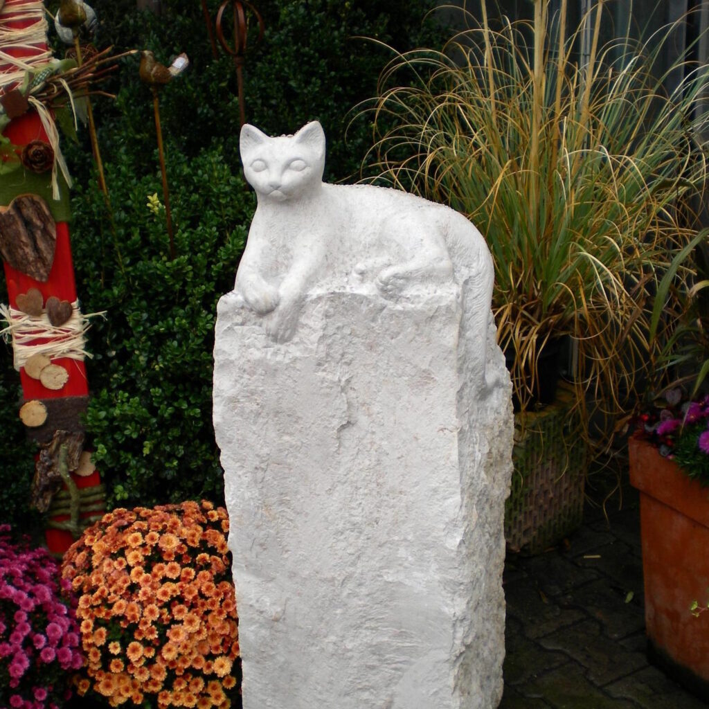 Katzenstatue auf einem Steinsockel zwischen Blumen. Dekorative Gartengestaltung. Steinmetz Meisterbetrieb