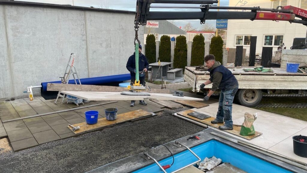 Handwerker verlegen Poolumrandung mit Kran. Pflasterarbeiten vom Meisterbetrieb. Steinmetz Meisterbetrieb