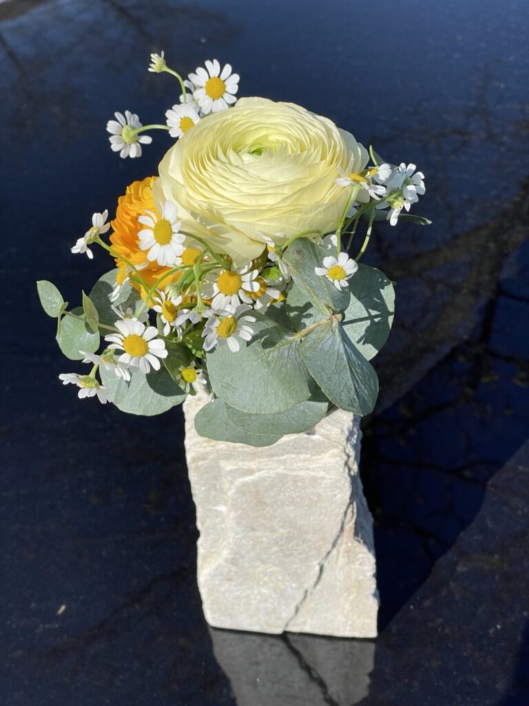 Steindekoration: Blumenarrangement mit Ranunkel, Gänseblümchen und Eukalyptus in einem Steinblock. Steinmetz Meisterbetrieb
